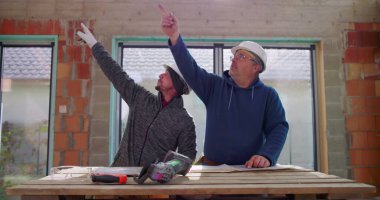 İnşaat işçileri inşaat alanındaki ahşap masa üzerinde öğütücü alet kullanıyorlar, takım çalışması ve işbirliği, güvenlik kaskları, yenileme projesi üzerine odaklanmış tartışmalar,