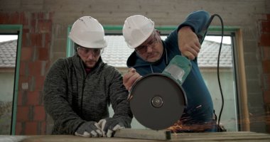 Metal üzerinde açı öğütücü kullanan odaklanmış inşaat işçileri, kıvılcımlar uçuşuyor, takım çalışması, ikisi de güvenlik kaskı ve koruyucu gözlük takıyor.