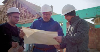 Mühendis ve inşaat işçileri inşaat alanının planlarını tartışıyor, kısmen inşa edilmiş yapı iskeleti önünde proje detaylarını gözden geçiriyorlar.