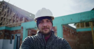 İnşaat alanında kolları bağlı duran erkek inşaat işçisi, gururlu bir profesyonel güven gösteriyor, inşaat endüstrisine bağlılık gösteriyor, arka planda proje var.