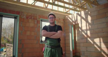 Genç inşaat işçisi kendine güvenerek kollarını kavuşturuyor. Güvenlik kaskı takıyor. İnşaat işlerinde doğal ışık, güç ve gururla tamamlanmamış inşaat sahasında duruyor.