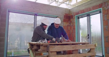 İki inşaat işçisi ahşap bir masanın üzerindeki bina planlarını gözden geçiriyorlar, içerideki yenileme projesini tartışıyorlar, yetenekli işçiler arasındaki işbirliği, güvenlik kaskları, takım çalışması