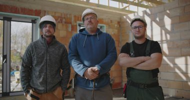 İçeride poz veren üç inşaat işçisi, tamamlanmamış inşaat sahasında güvenlik kaskı takıyorlar, inşaat ortamında profesyoneller arasında takım çalışması ve dostluk.