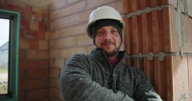 Gülümseyen inşaat işçisi kollarını kavuşturuyor, miğfer ve gri ceket giyiyor, tamamlanmamış bir binada duruyor, tuğla duvara yaslanıyor, kendinden emin ve cana yakın.