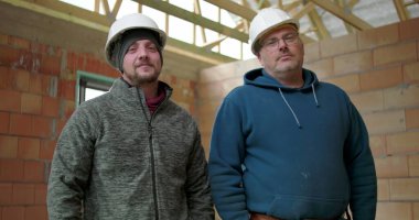 Kapalı alanda duran inşaat işçileri, kendine güvenen pozlar, inşaat alanında tamamlanmamış yapıda iki adam, takım ruhu ve inşaat ortamında iş gururu.