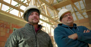 Bitmemiş bir binanın içinde iki inşaat işçisinin düşük açılı görüntüsü, kolları çapraz, kendinden emin ve iddialı, somut takım çalışması ve profesyonellik.