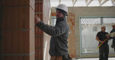 Duvarı kalemle ölçen inşaat işçisi, güvenlik miğferinde odaklanmış miğferle içeride tuğla yapı, hassasiyet ve detaylara dikkat ediyor.