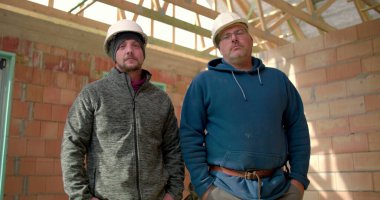 İnşaat alanında kapalı alanda poz veren iki inşaatçı, güvenlik kasklı adamlar takım çalışması ve güven gösteriyor, tamamlanmamış tuğla yapıda çalışıyorlar, ilerleme kaydediyorlar.