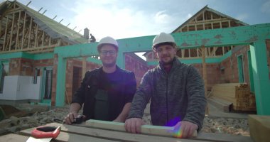 Yeni ev inşaatında iki erkek inşaatçı aletlerle ayakta duruyor, kameraya bakıyor, başlık takıyor, konut projesinde çalışıyor, çerçeveyi görünür kılıyor.