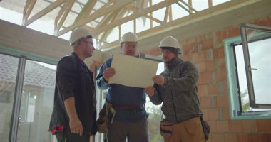 İnşaat ekibi inşaat planlarını analiz ediyor ve mimari detaylara işaret ediyor. Proje koordinasyonu için tamamlanmamış bir yapının içinde işbirliği yapıyorlar.