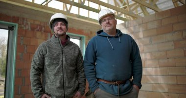 İnşaat alanında poz veren iki inşaat işçisi, güvenlik kaskı takan adamlar tamamlanmamış yapıda yan yana duruyorlar, takım arkadaşlığı ve iş gururu.