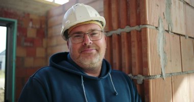 İnşaat işçisi tamamlanmamış binanın içinde duruyor, tuğla duvara yaslanıyor, güvenlik kaskı ve gözlük takıyor, kollar çapraz, kendinden emin ve cana yakın.