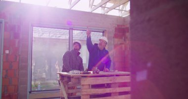 İnşaat işçileri inşaat alanındaki ahşap masa üzerinde öğütücü alet kullanıyorlar, takım çalışması ve işbirliği, güvenlik kaskları, yenileme projesi üzerine odaklanmış tartışmalar,