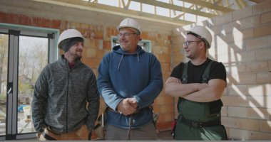 Üç inşaat işçisi kapalı alanda birlikte duruyorlar, güvenlik kaskı takıyorlar, bitmemiş bir inşaat sahasında gülümsüyorlar ve birlik gösteriyorlar, takım çalışmasını ve arkadaşlığı temsil ediyorlar.