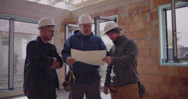 Üç inşaat işçisi planlarını gözden geçiriyorlar, güvenlik kaskı takıyorlar, proje detaylarını tartışıyorlar, takım çalışması ve bitmemiş endüstriyel alanda işbirliği yapıyorlar.