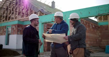 Dış inşaat alanının planlarını inceleyen üç inşaat işçisinden oluşan bir grup, kısmen inşa edilmiş binanın önündeki bina planlarını tartışıyor.
