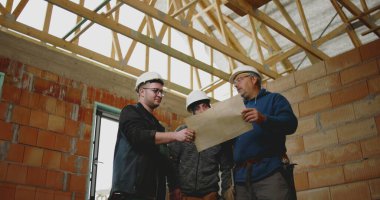 İnşaat ekibi inşaat planlarını analiz ediyor ve mimari detaylara işaret ediyor. Proje koordinasyonu için tamamlanmamış bir yapının içinde işbirliği yapıyorlar.