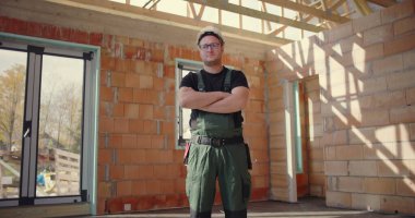 İnşaat işçisi güvenle duruyor. Kolları bağlı, kaskı ve tulumuyla inşaat alanında duruyor. İnşaat alanında gururlu ve profesyonel duruş sergiliyor.