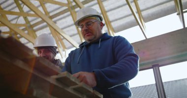 Tahta tutan inşaat işçisinin düşük açılı görüntüsü, yapısal projede takım çalışması, koruyucu kask, odaklı ifade, görünür yapı iskeleti.