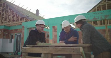 İnşaat alanında işbirliği yapan üç inşaat işçisi, bitmemiş yapının yakınındaki ahşap işleme programının proje detaylarını tartışıyorlar.