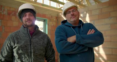 İki inşaat işçisi kısmen inşa edilmiş bir yapının içinde dikiliyorlar, başlık takıyorlar, birinin kolları çaprazlanmış, yoldaşlık ve iş yerinde birlik sağlıyorlar.