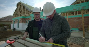 Bir inşaat sahasında ahşabı ölçüp işaretleyen iki müteahhit, takım çalışması ve hassasiyet, konut projesi, yeni bir ev, sağlam şapkalar, işe yarar araçlar.