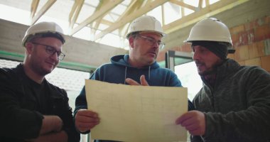 İnşaat ekibi tamamlanmamış binanın planlarını inceliyor, proje detayları, güvenlik kaskları, sanayi ortamında takım çalışması ve planlama üzerine odaklanmış tartışmalar yapıyor