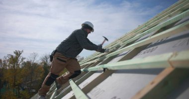 İnşaat alanında çatı iskeletini çekiçle güvence altına alan işçiler, güvenlik kaskı takıyorlar, çatıdaki hareketliliğin yan görüntüsü, açık hava inşaat projesinin bir parçası.