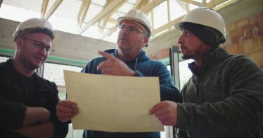 İnşaat ekibi tamamlanmamış binanın planlarını inceliyor, proje detayları, güvenlik kaskları, sanayi ortamında takım çalışması ve planlama üzerine odaklanmış tartışmalar yapıyor