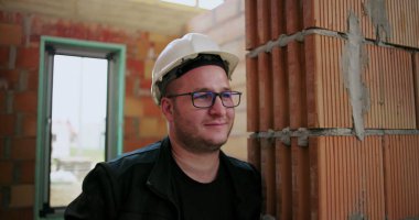 Kask ve gözlük takan, inşaat halindeki tuğladan duvara yaslanan, rahat ve dost canlısı tavırlar sergileyen gülümseyen inşaat işçilerinin yakın çekimi.