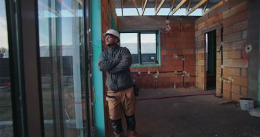 Genç inşaat işçisi, cam baret takılı, içinde inşaat malzemeleri ve açık tavan kirişleriyle dışarıda gözetleme yapıyor.