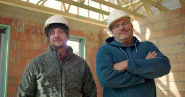 İnşaat halindeki bir binada yan yana duran iki inşaat işçisi, biri gülümsüyor diğeri ise kollarını kavuşturup takım çalışması ve çalışma arkadaşlığı yapıyor.