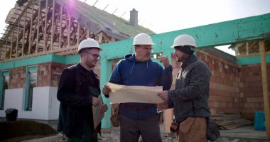 Dış inşaat alanının planlarını inceleyen üç inşaat işçisinden oluşan bir grup, kısmen inşa edilmiş binanın önündeki bina planlarını tartışıyor.