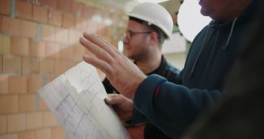 İnşaat planlarını, miğferleri, takım çalışmasını ve işbirliğini tartışan inşaat işçilerinin ayrıntılı görüşü, plan üzerinde yoğunlaşmak, inşaat alanı