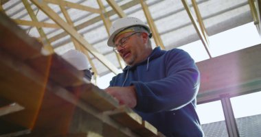 Tahta tutan inşaat işçisinin düşük açılı görüntüsü, yapısal projede takım çalışması, koruyucu kask, odaklı ifade, görünür yapı iskeleti.