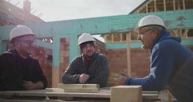 Üç inşaat işçisi inşaat alanındaki planları tartışıyor, başlık takıyor, çalışma masasında duruyor, sohbet ediyor, konut inşaatı projesiyle uğraşıyor.