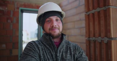 Kolları bağlı inşaat işçisi, güvenlik kaskı takılı, tamamlanmamış bina içinde duruyor, kameraya bakıyor, endüstriyel ayarlar, profesyonel davranışlar.