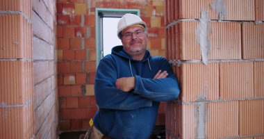 İnşaat işçisi inşaat halindeki tuğladan duvara yaslanıyor, kollar çapraz, güvenlik kaskı takıyor, rahat ve kendinden emin duruş, endüstriyel ortam.