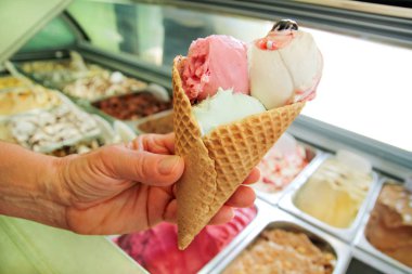 Kadın elini büyük bir çilek, nane, vişne dondurma waffle koni ile vanilya tutuyor. Dondurma buzdolabı çelik hizmet kapsayıcıları içinde geçmiş. Dondurma dükkanı işçi. Pastane.