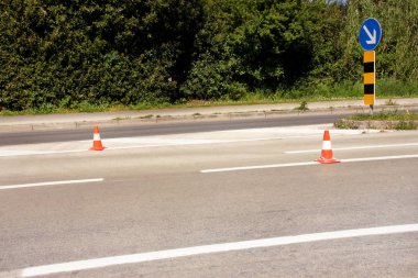 Yolda çalış. Trafik işaretli inşaat konileri sağdaki tabelayı tutar. Asfalt üzerinde beyaz ve turuncu çizgileri olan trafik konileri. Trafik işaretleri ve yol bakımı, inşaat tabelası altında..