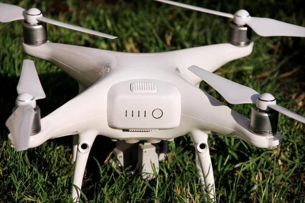 White quadcopter Drone with 4K digital camera on grass is ready for take off to fly in air to take photos, record footage from above. Drone with four motors, propellers, camera and red warning lights.