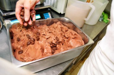 Şef üniforma içinde kadın pudra dondurma fabrikasında çalışıyor. O karanlık çikolatalı dondurma lezzeti ve çelik kapsayıcı ekleme çikolata crunch bisküvi parçaları süslemeleri. Üretim.