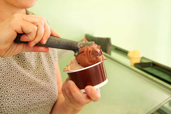 Female hand with scoop takes ice cream from the fridge and serving in cardboard ice cream cup. Woman taking scoop of tasty ice cream. Fridge with ice cream. Woman works in ice cream shop. Pastry shop.