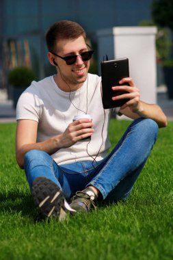 Tablet ve kulaklık kullanarak güneş gözlüklü genç yakışıklı adam, kahve içme çimenlerin üzerinde oturan ve güzel bir güneşli gün modern bina tarafından zevk. Erkek model çekimi, kentsel moda yaşam tarzı.