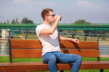 Yakışıklı çocuk bir iskelede bir bankta oturmuş ve kahve içme güneş gözlüğü takıyor. Şehir rush bir mola. Arka plan sahne Köprüsü, nehir, iskele, yol nehir ve yeşil çimen.