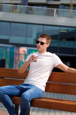 Adam bankta oturan, sabah kahve içiyor. Şehir rush mola, poz ve güneşli bir gün zevk. Modern binalar ve çağdaş mimari ayrıntılı arka plan olarak kentsel sahnesi.