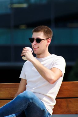 Adam bankta oturan, sabah kahve içiyor. Şehir rush mola, poz ve güneşli bir gün zevk. Modern binalar ve çağdaş mimari ayrıntılı arka plan olarak kentsel sahnesi.