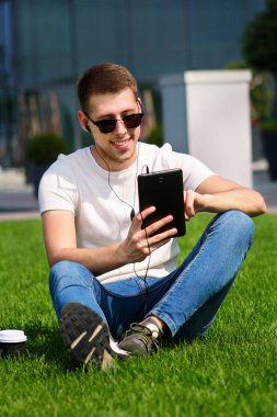 Tablet ve kulaklık kullanarak güneş gözlüklü genç yakışıklı adam, kahve içme çimenlerin üzerinde oturan ve güzel bir güneşli gün modern bina tarafından zevk. Erkek model çekimi, kentsel moda yaşam tarzı.