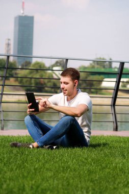 Yakışıklı çocuk tablet ve kulaklık kullanarak, kahve içme dostum çimenlerin üzerinde oturan ve nehir yanında güzel bir güneşli gün zevk. Model fotoğraf çekimi, kentsel moda, yaşam tarzı. Beyaz T-shirt ve jeans.