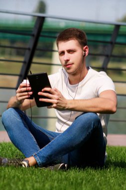 Yakışıklı çocuk tablet ve kulaklık kullanarak, kahve içme dostum çimenlerin üzerinde oturan ve nehir yanında güzel bir güneşli gün zevk. Model fotoğraf çekimi, kentsel moda, yaşam tarzı. Beyaz T-shirt ve jeans.